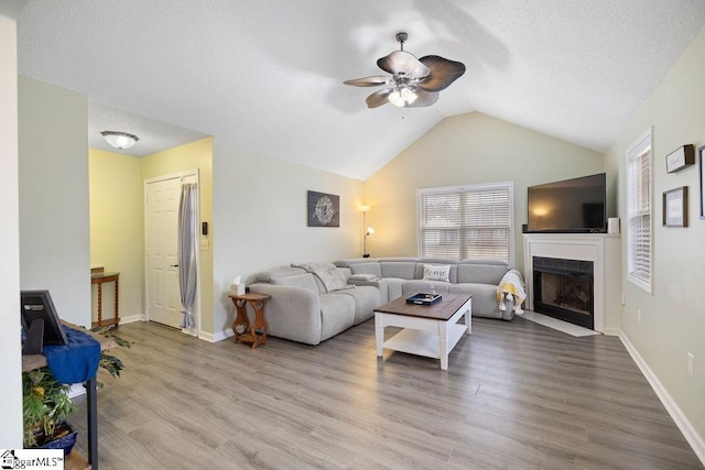 living area featuring ceiling fan, lofted ceiling, a fireplace with flush hearth, wood finished floors, and baseboards