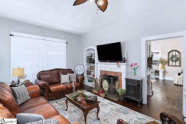 living room with baseboards, dark wood finished floors, ceiling fan, a brick fireplace, and built in shelves