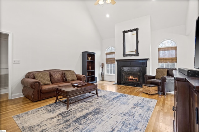 living room with a warm lit fireplace, high vaulted ceiling, light wood-style flooring, and a ceiling fan