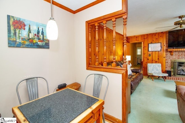 dining room featuring wooden walls, carpet floors, a ceiling fan, ornamental molding, and a brick fireplace