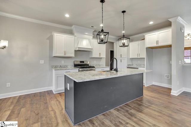 kitchen featuring an island with sink, custom exhaust hood, stainless steel range with gas cooktop, white cabinetry, and a sink