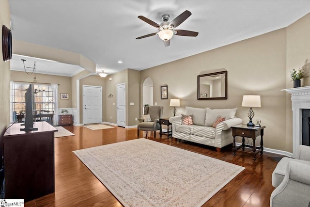 living area featuring arched walkways, ceiling fan with notable chandelier, a fireplace, baseboards, and dark wood finished floors