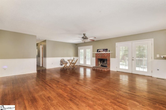 unfurnished living room featuring a wainscoted wall, ceiling fan, wood finished floors, french doors, and a brick fireplace