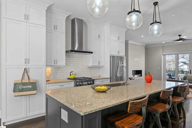 kitchen featuring pendant lighting, wall chimney range hood, an island with sink, and white cabinets