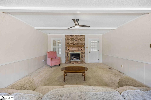 living room featuring beam ceiling, a brick fireplace, wainscoting, ceiling fan, and light carpet