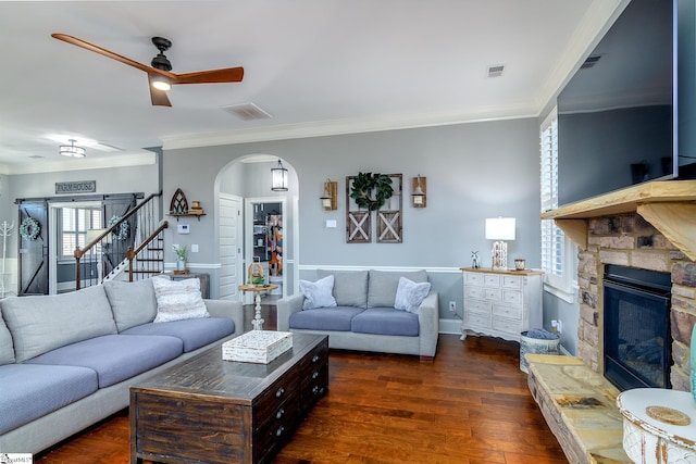 living room with arched walkways, a stone fireplace, stairs, ornamental molding, and dark wood-style floors