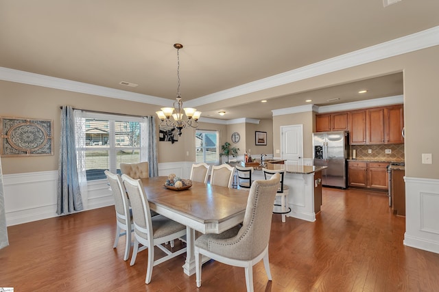 dining space with a wainscoted wall, dark wood-style flooring, an inviting chandelier, crown molding, and a decorative wall