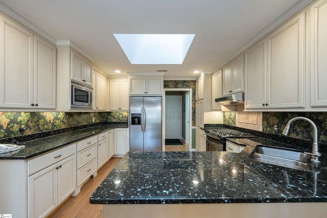 kitchen with a skylight, appliances with stainless steel finishes, dark stone countertops, under cabinet range hood, and a sink