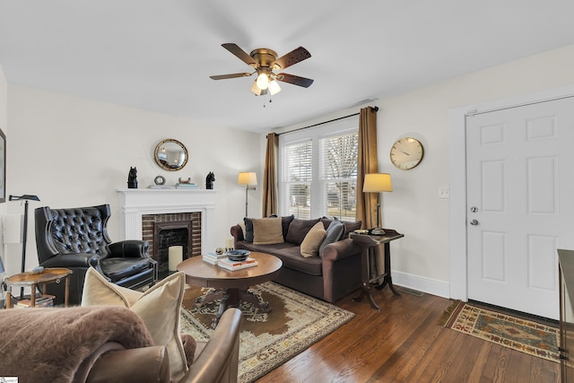 living area with dark wood-type flooring, a brick fireplace, a ceiling fan, and baseboards