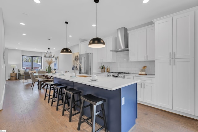 kitchen with hanging light fixtures, wall chimney range hood, an island with sink, and white cabinetry