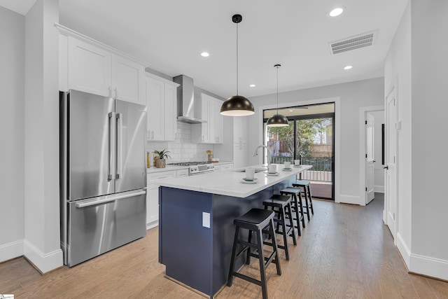 kitchen with stainless steel appliances, white cabinetry, light countertops, wall chimney range hood, and a center island with sink