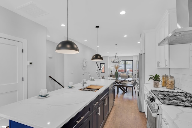 kitchen featuring decorative light fixtures, a center island with sink, stainless steel appliances, white cabinets, and wall chimney range hood