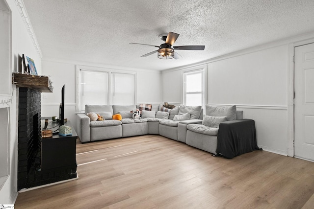 living room featuring a brick fireplace, ceiling fan, light wood-style flooring, and a textured ceiling