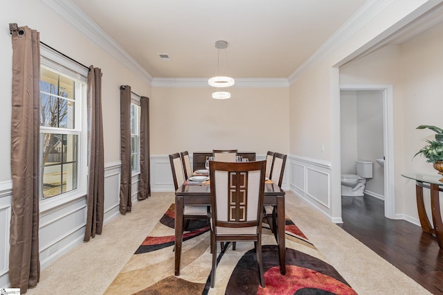 carpeted dining area with ornamental molding, wainscoting, and a decorative wall