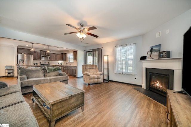 living area featuring a glass covered fireplace, baseboards, ceiling fan, and light wood finished floors