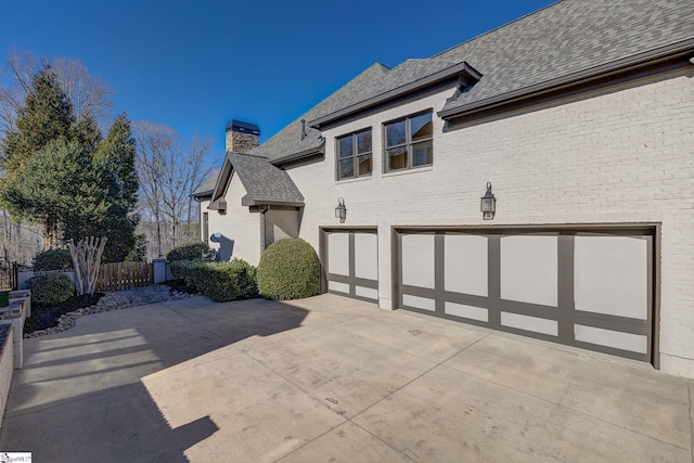 view of side of property with a garage, brick siding, concrete driveway, roof with shingles, and a chimney