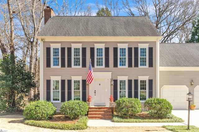 colonial house with a garage, concrete driveway, a shingled roof, and a chimney