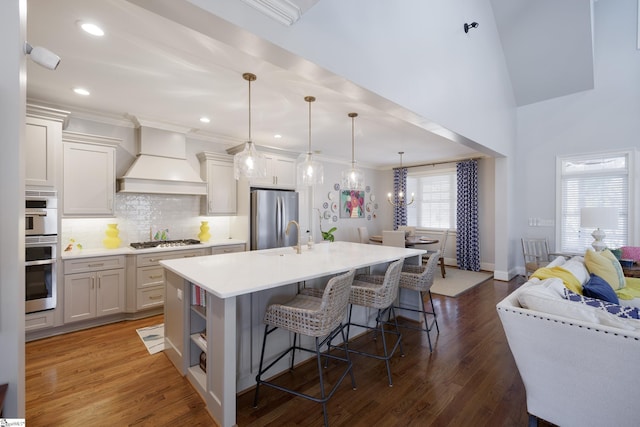 kitchen featuring an island with sink, custom range hood, decorative light fixtures, stainless steel appliances, and light countertops