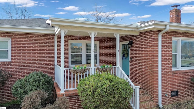 view of exterior entry with covered porch, brick siding, a shingled roof, crawl space, and a chimney