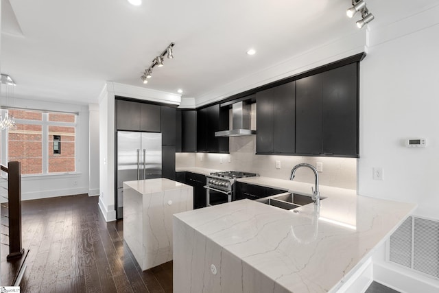 kitchen with a peninsula, a sink, high quality appliances, wall chimney range hood, and dark cabinetry