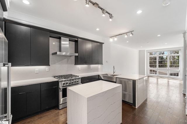 kitchen featuring appliances with stainless steel finishes, a sink, dark cabinetry, a peninsula, and wall chimney exhaust hood