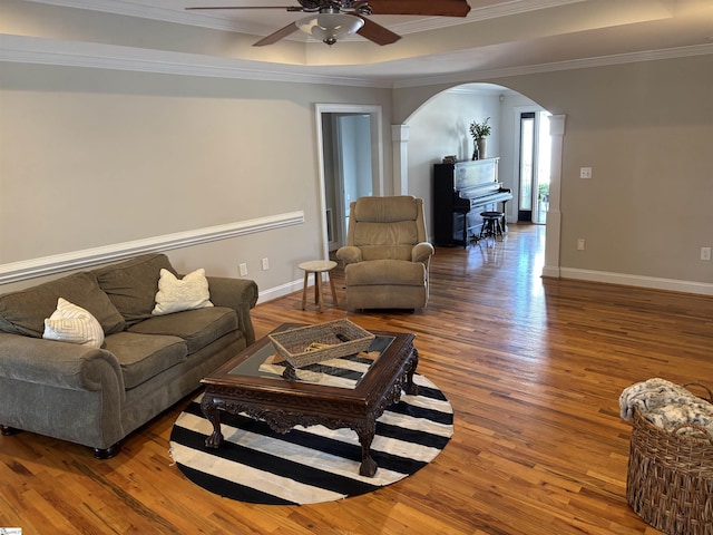 living area with arched walkways, wood finished floors, baseboards, a tray ceiling, and crown molding