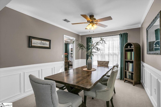 dining area with light colored carpet, a wainscoted wall, a ceiling fan, visible vents, and crown molding