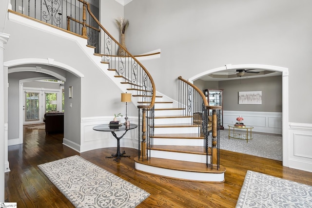 stairs with arched walkways, ceiling fan, a high ceiling, wood finished floors, and wainscoting