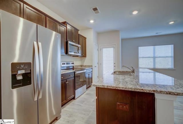 kitchen featuring light stone counters, light wood finished floors, stainless steel appliances, a sink, and an island with sink