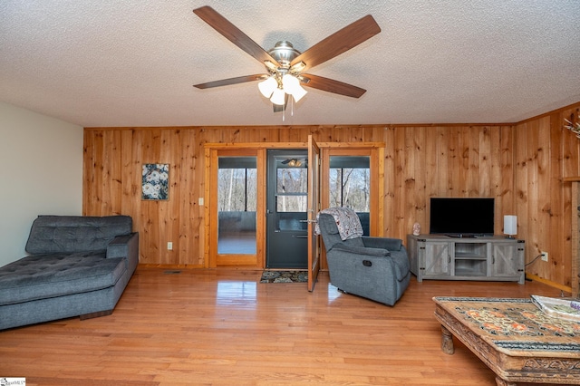 living area featuring light wood-style flooring, wooden walls, a ceiling fan, and a textured ceiling