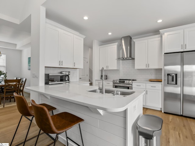 kitchen with a breakfast bar area, appliances with stainless steel finishes, light wood-style floors, white cabinets, and wall chimney range hood