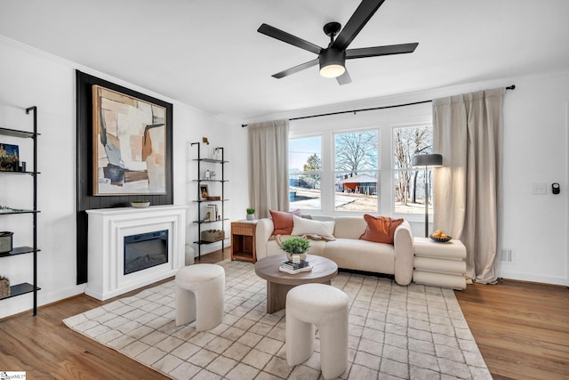 living area featuring ceiling fan, baseboards, light wood finished floors, a glass covered fireplace, and crown molding