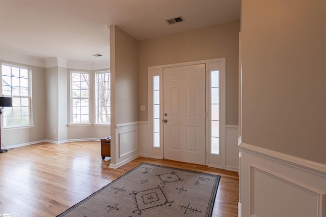 foyer entrance featuring light wood-type flooring, a wainscoted wall, visible vents, and crown molding