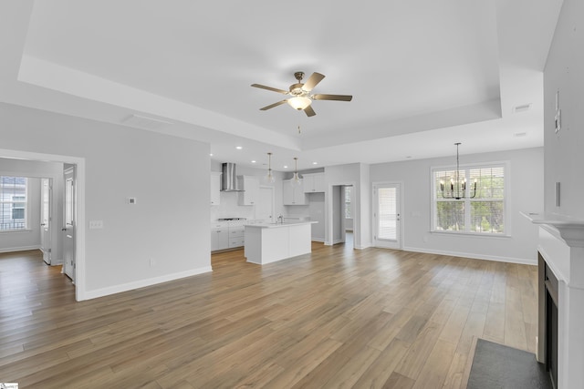 unfurnished living room with light wood-style flooring, baseboards, a raised ceiling, and ceiling fan with notable chandelier