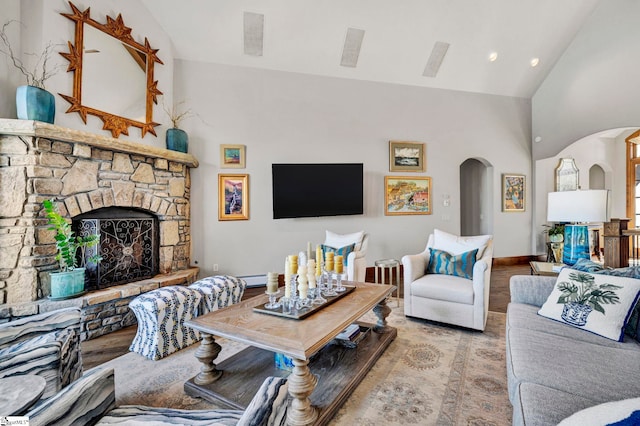 living area featuring baseboards, arched walkways, wood finished floors, a stone fireplace, and high vaulted ceiling