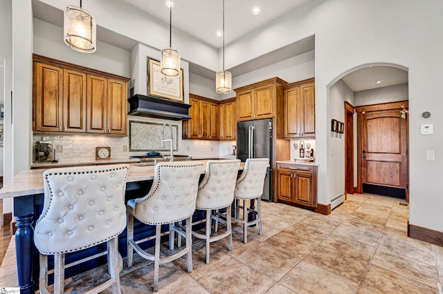 kitchen with arched walkways, backsplash, freestanding refrigerator, brown cabinets, and an island with sink