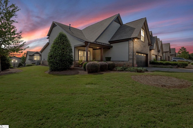 property exterior at dusk with a garage, brick siding, a shingled roof, concrete driveway, and a lawn