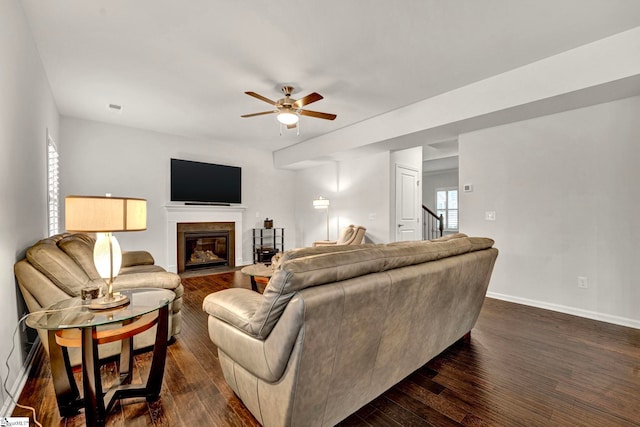 living room featuring dark wood-type flooring, a fireplace with flush hearth, a ceiling fan, baseboards, and stairs