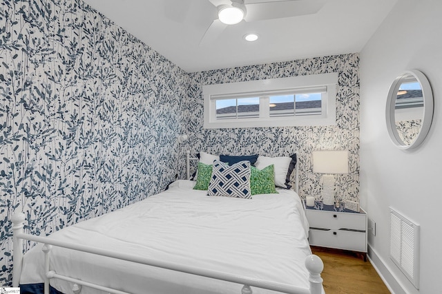 bedroom featuring visible vents, a ceiling fan, wood finished floors, baseboards, and wallpapered walls