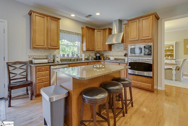 kitchen with stainless steel appliances, a sink, a kitchen island, light wood-type flooring, and wall chimney exhaust hood