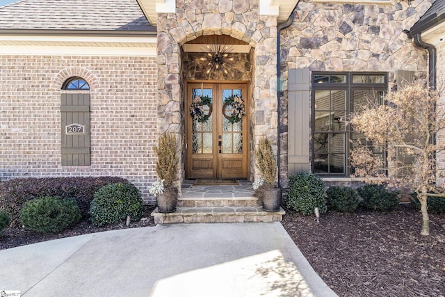 entrance to property featuring stone siding, brick siding, roof with shingles, and french doors