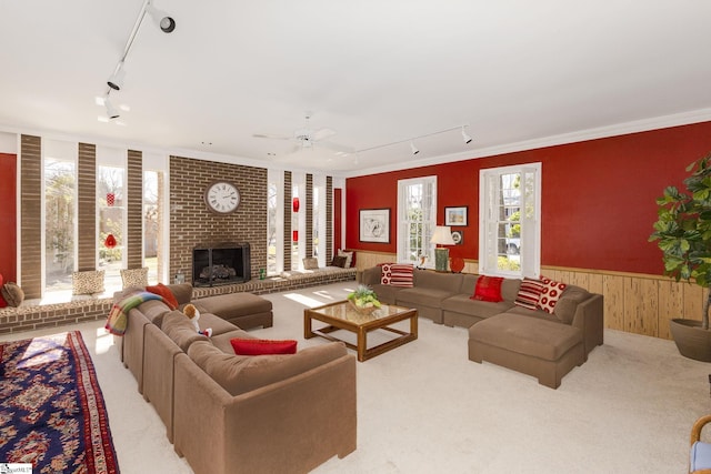 carpeted living area featuring ceiling fan, wood walls, wainscoting, a brick fireplace, and track lighting