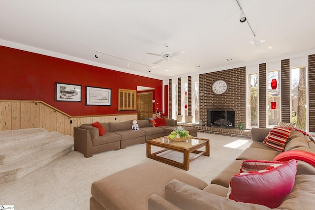 carpeted living area featuring a wainscoted wall, rail lighting, ornamental molding, a brick fireplace, and ceiling fan