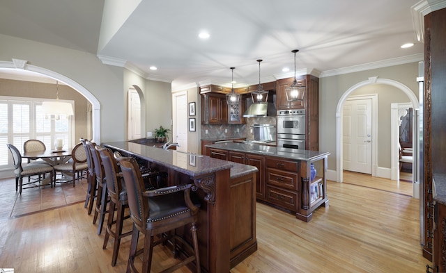 kitchen featuring arched walkways, decorative backsplash, double oven, a kitchen island with sink, and wall chimney range hood