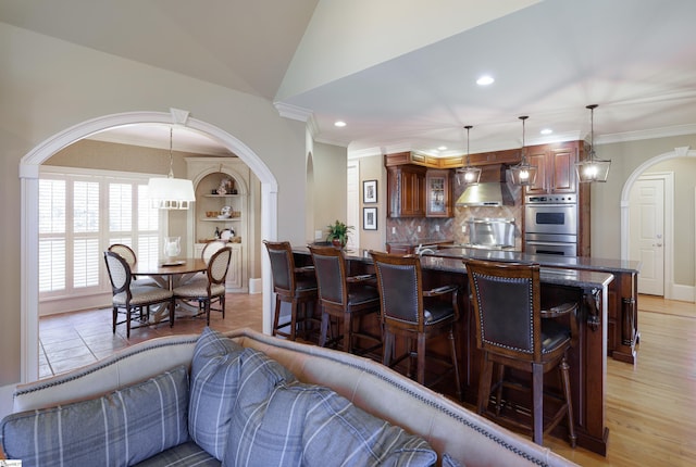 interior space with arched walkways, a breakfast bar area, decorative backsplash, ornamental molding, and wall chimney exhaust hood