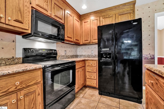 kitchen with wallpapered walls, brown cabinetry, light stone countertops, black appliances, and light tile patterned flooring