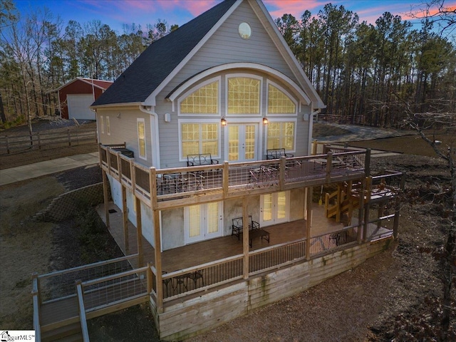 back of property at dusk featuring a garage, a shingled roof, french doors, a wooden deck, and an outdoor structure