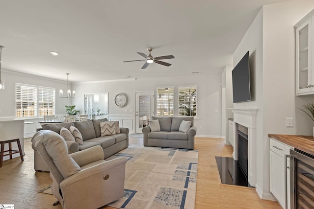 living room featuring baseboards, a fireplace with flush hearth, wine cooler, light wood-type flooring, and ceiling fan with notable chandelier