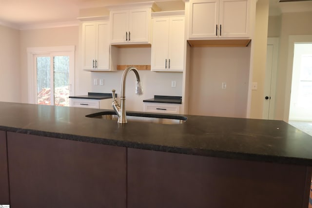 kitchen with crown molding, a center island with sink, dark countertops, white cabinetry, and a sink