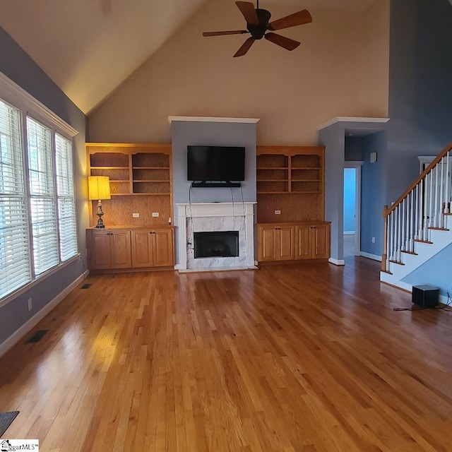 unfurnished living room featuring high vaulted ceiling, a premium fireplace, a ceiling fan, stairs, and light wood-type flooring